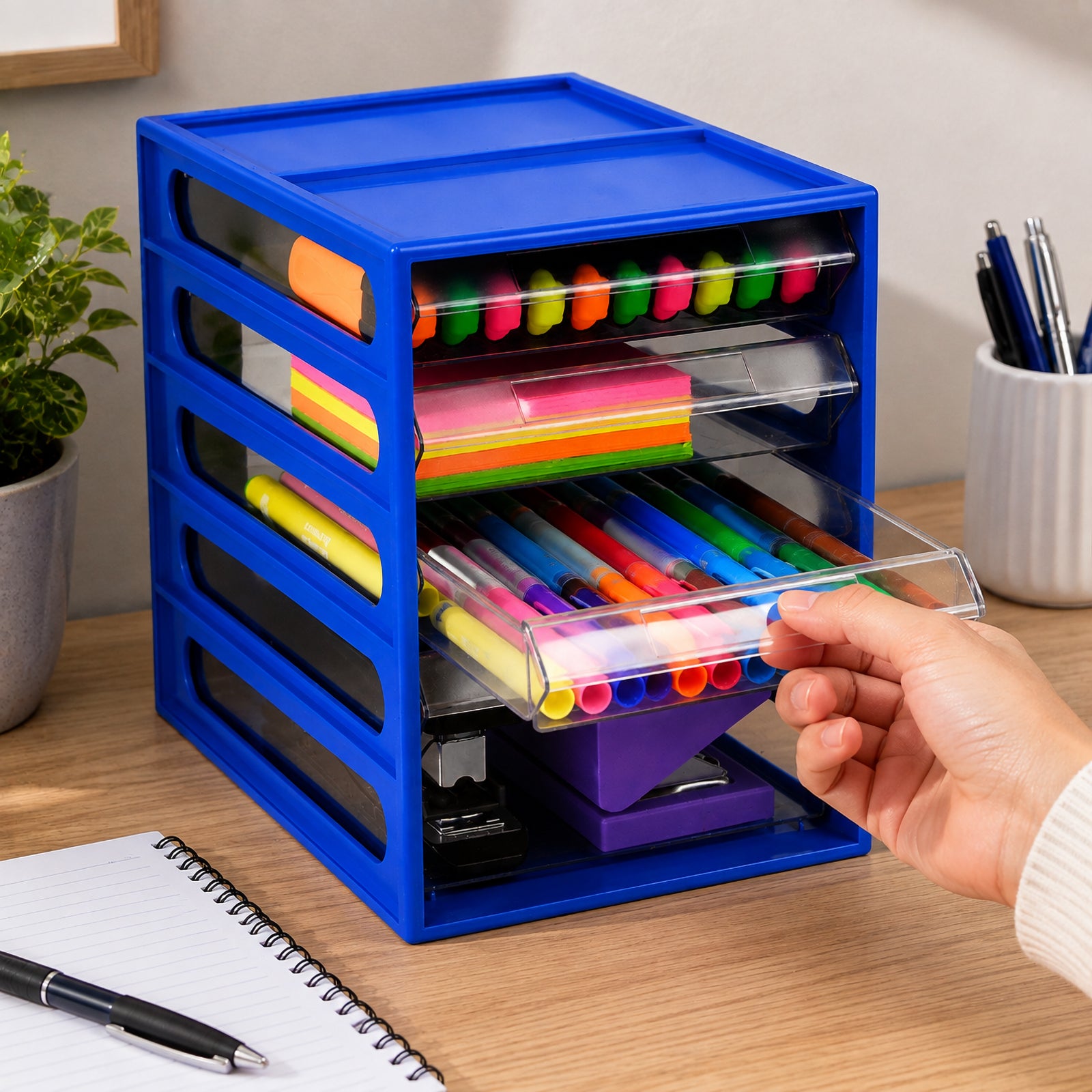 Italplast 4 Drawer Office Organiser in Blueberry displayed on a timber desk beside a notebook and pen. A hand opens one of the clear drawers containing colourful writing tools, while highlighters and sticky notes are stored above and larger stapling equipment sits below, demonstrating convenient access to organised everyday stationery.