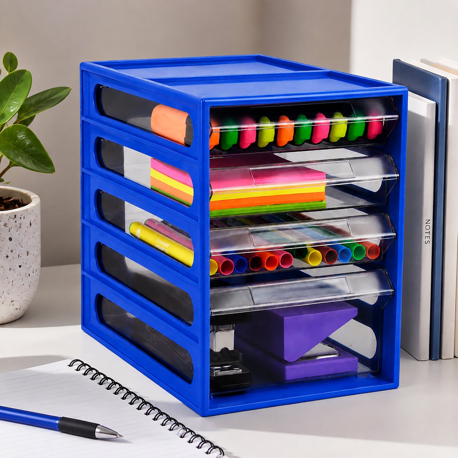 Angled lifestyle view of the Italplast 4 Drawer Office Organiser in Blueberry positioned beside notebooks and a plant. Clear drawers display colourful stationery across the upper compartments with larger desktop tools stored in the lower section, highlighting the compact vertical design for organised offices, study areas and administration workspaces.