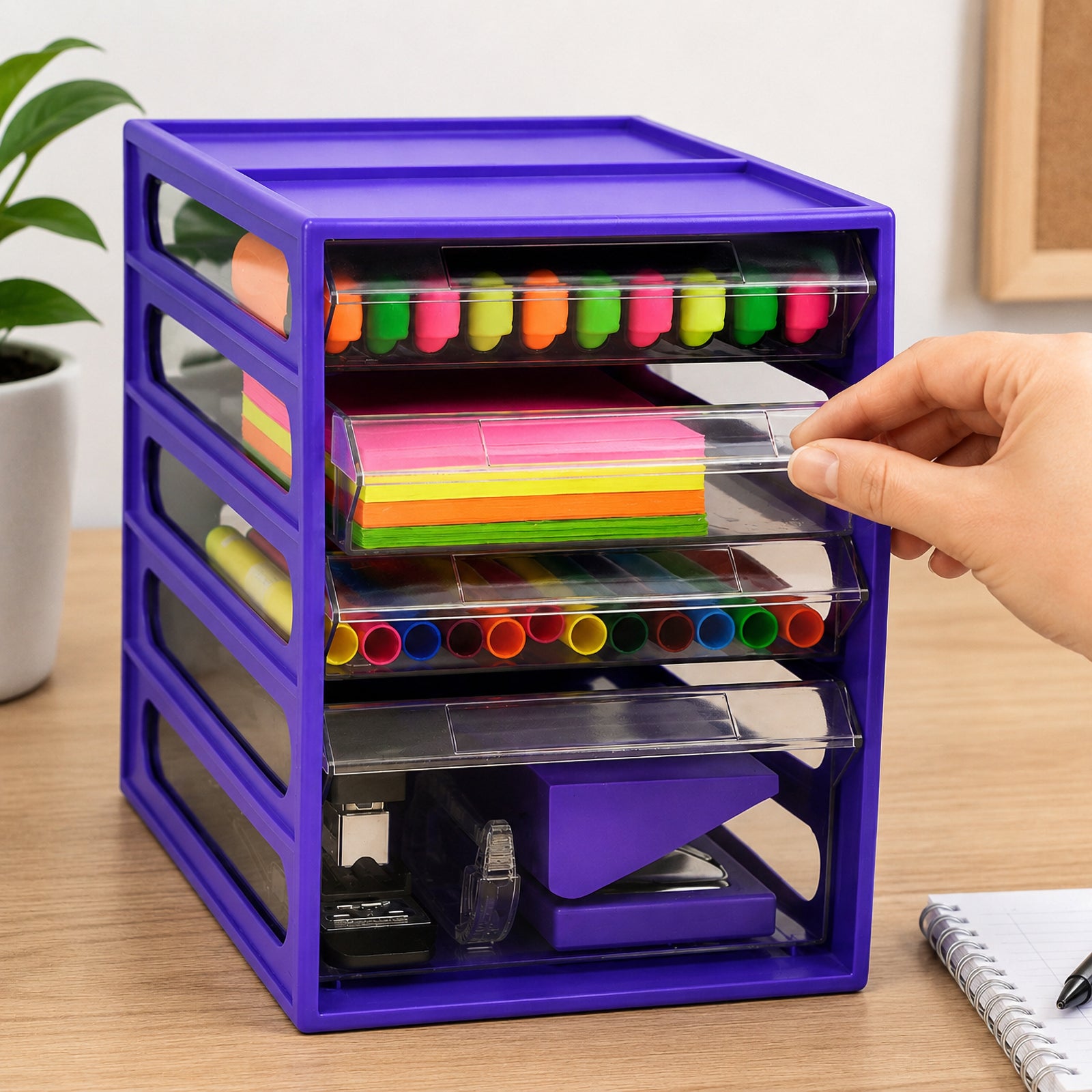 Italplast 4 Drawer Office Organiser in Grape on a timber desk, with hands opening the spacious clear lower drawer containing stapling and tape equipment. Three smaller transparent drawers above organise colourful highlighters, sticky notes and writing supplies, demonstrating accessible stationery storage beside a notebook and everyday office accessories.