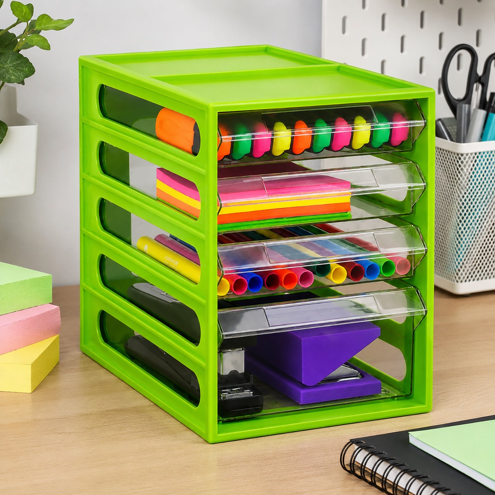 Angled lifestyle view of the Italplast 4 Drawer Office Organiser in Lime on an organised workspace. Three smaller clear drawers separate colourful stationery and writing supplies, while the deeper lower drawer accommodates larger desktop tools. The compact upright format demonstrates practical vertical storage for keeping frequently used supplies accessible.