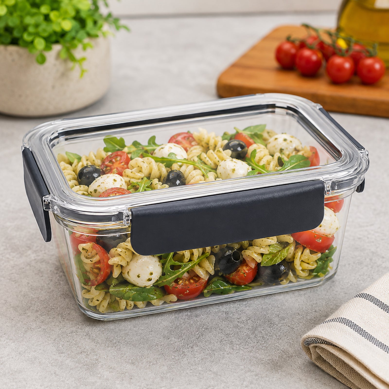 Angled view of the Italplast 1300ml Snap Lock Food Container filled with pasta salad, tomatoes, olives, greens and cheese. Displayed on a kitchen benchtop, the image demonstrates the containers useful depth for individual meals while clearly showing its transparent rectangular construction, secure lid and dark snap lock closures around the edges.
