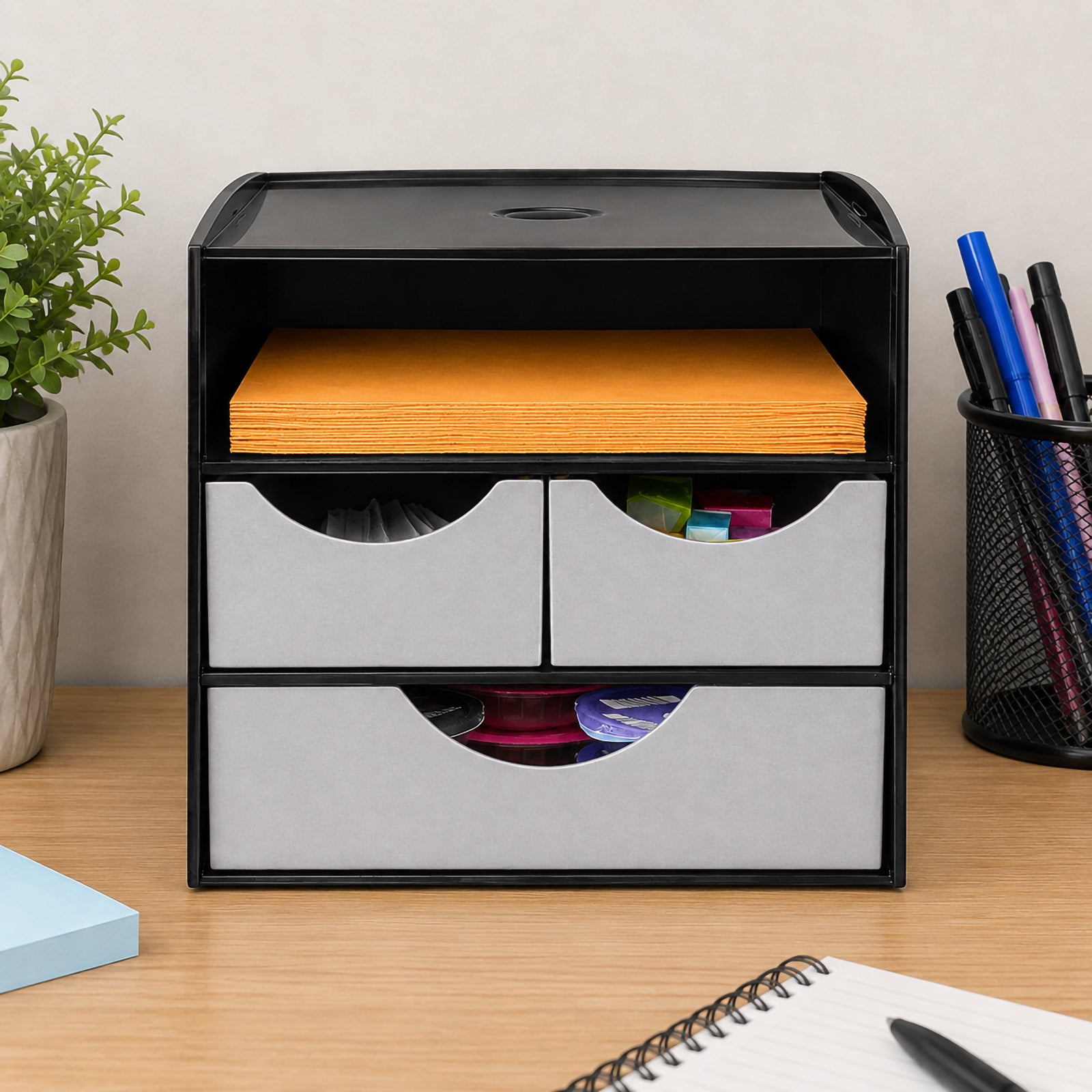 Front view of the Italplast Take A Break drawer organiser showing its compact black housing, two metallic grey upper drawers and one wide lower drawer. An open compartment above the drawers holds a stack of orange napkins, while small supplies are visible inside the drawer sections. The image demonstrates how the module can create a neat workplace refreshment or stationery station.