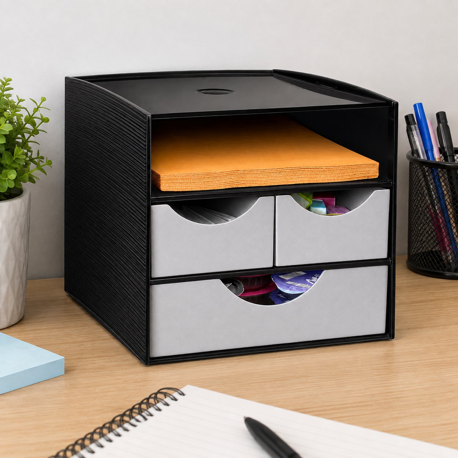 Angled view of the Italplast Take A Break 3 Tier Drawer Module arranged on an office desk beside a pen holder and notebook. The open upper shelf contains orange napkins, while the metallic grey drawers store assorted small supplies. The compact vertical design shows how multiple storage areas can help reduce benchtop clutter without occupying excessive desk space.