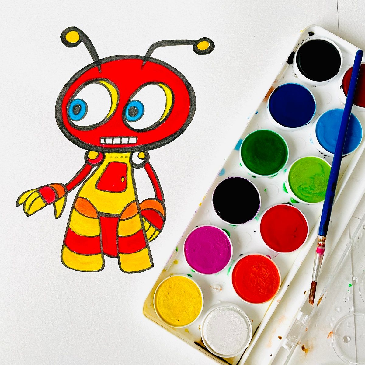Jasart Byron watercolour discs in use beside a brightly painted red and yellow cartoon bug drawing, with a brush resting in the palette