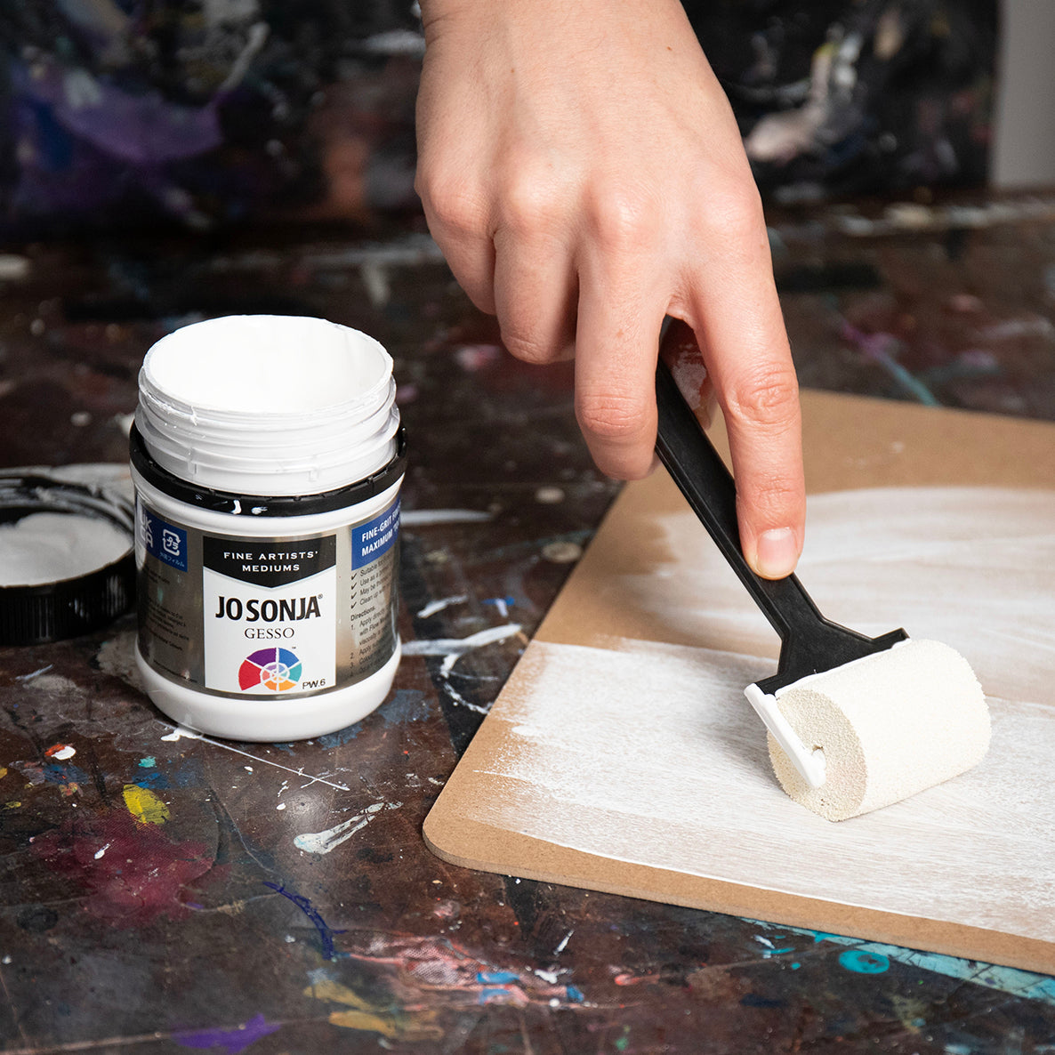 Hand rolling white Jo Sonja gesso onto a brown board with a foam roller beside an open jar of gesso on a paint-splashed bench