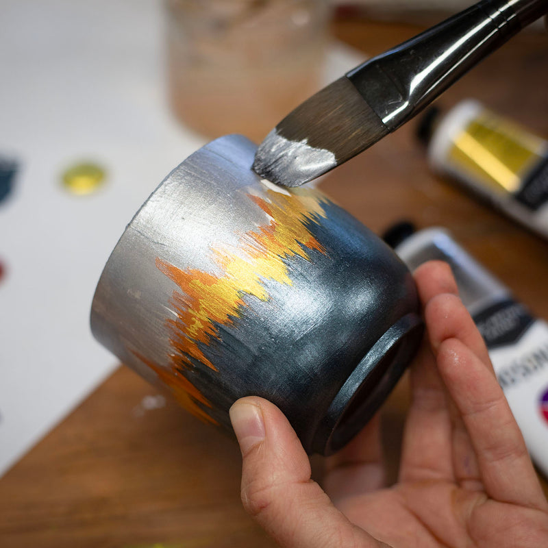 Hand holding a pot painted silver and black with gold flames while a flat brush adds silver