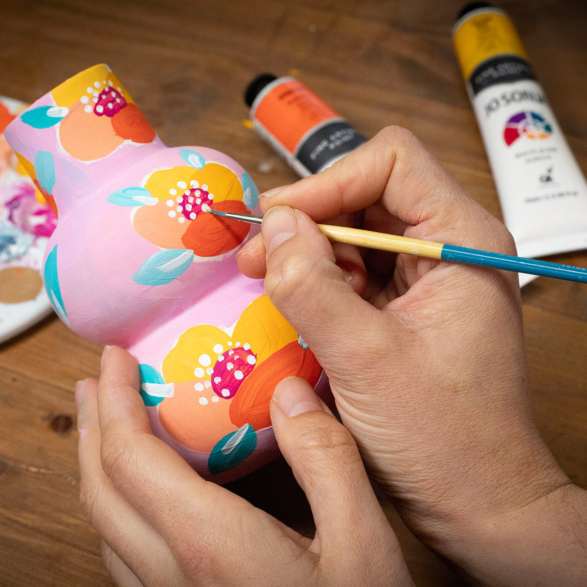 Hands painting a floral pattern on a pink ceramic vessel with Jo Sonja tubes on the bench