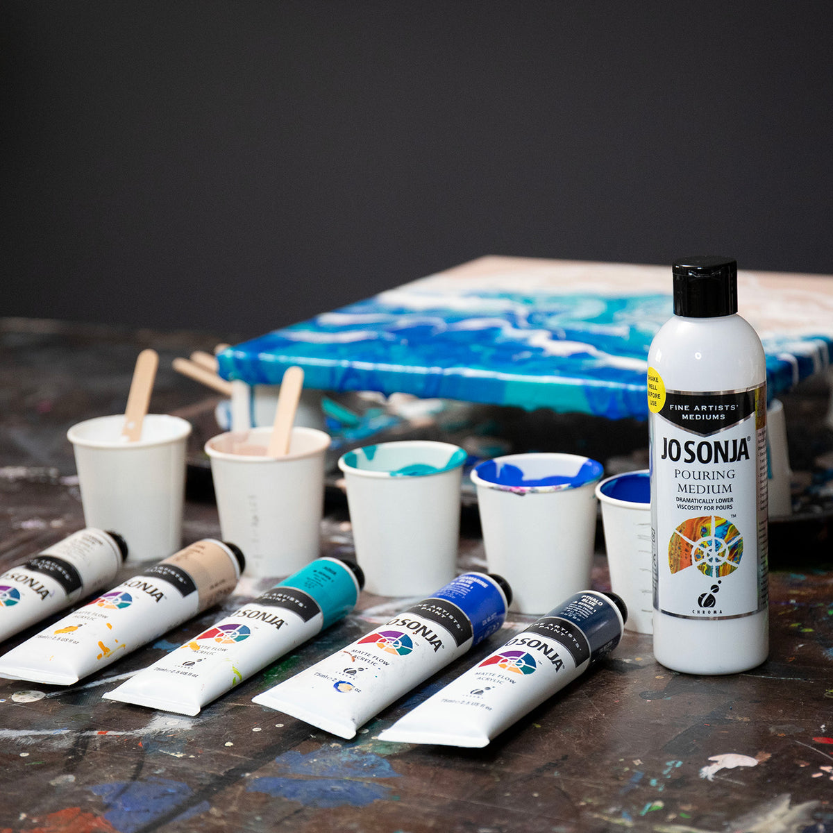 Jo Sonja Pouring Medium bottle on a paint-splashed table with cups of mixed colour, Jo Sonja paint tubes and a blue and white poured canvas
