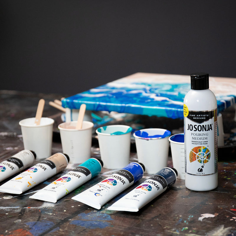 Jo Sonja Pouring Medium bottle on a paint-splashed table with cups of mixed colour, Jo Sonja paint tubes and a blue and white poured canvas