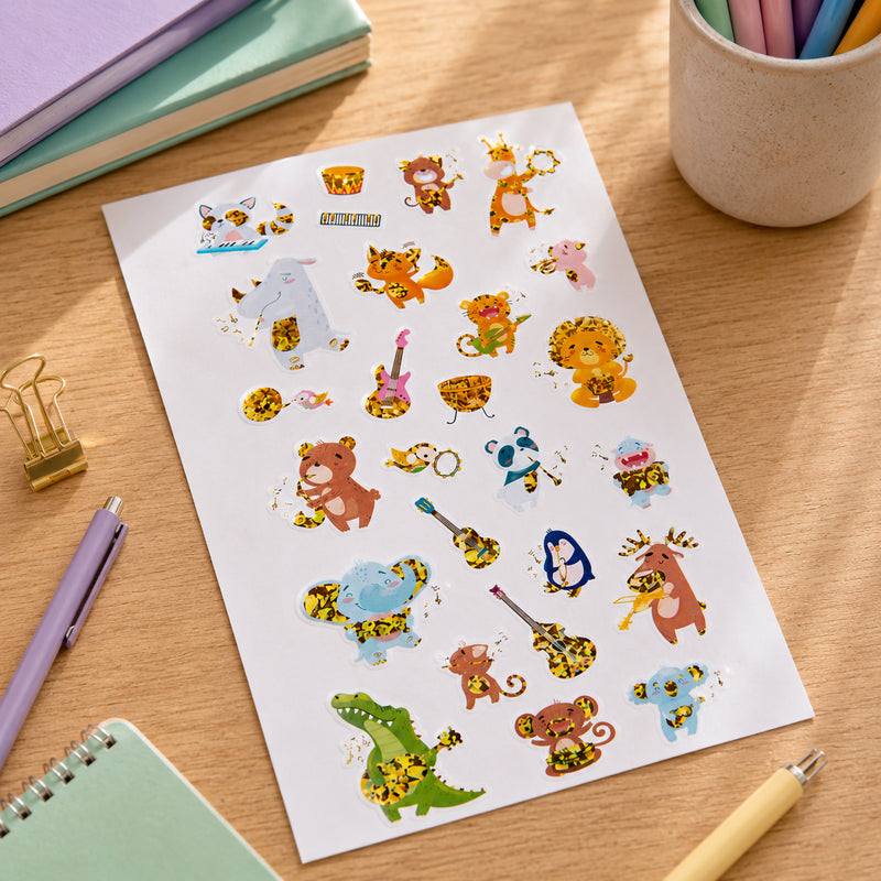 Close up of the full sticker sheet featuring cheerful jungle animals playing musical instruments including guitars, keyboards, tambourines and drums. The holographic foil accents create bright shimmering highlights throughout the design.
