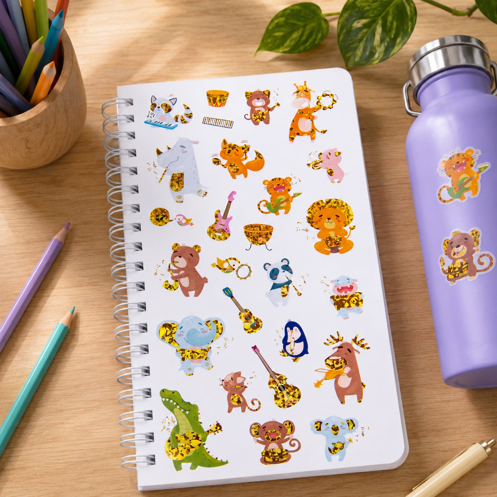 Spiral notebook decorated with colourful holographic jungle animal musician stickers including lions, tigers, monkeys, crocodiles, elephants and bears playing guitars, drums and percussion instruments. A purple drink bottle nearby also features the playful music themed stickers.