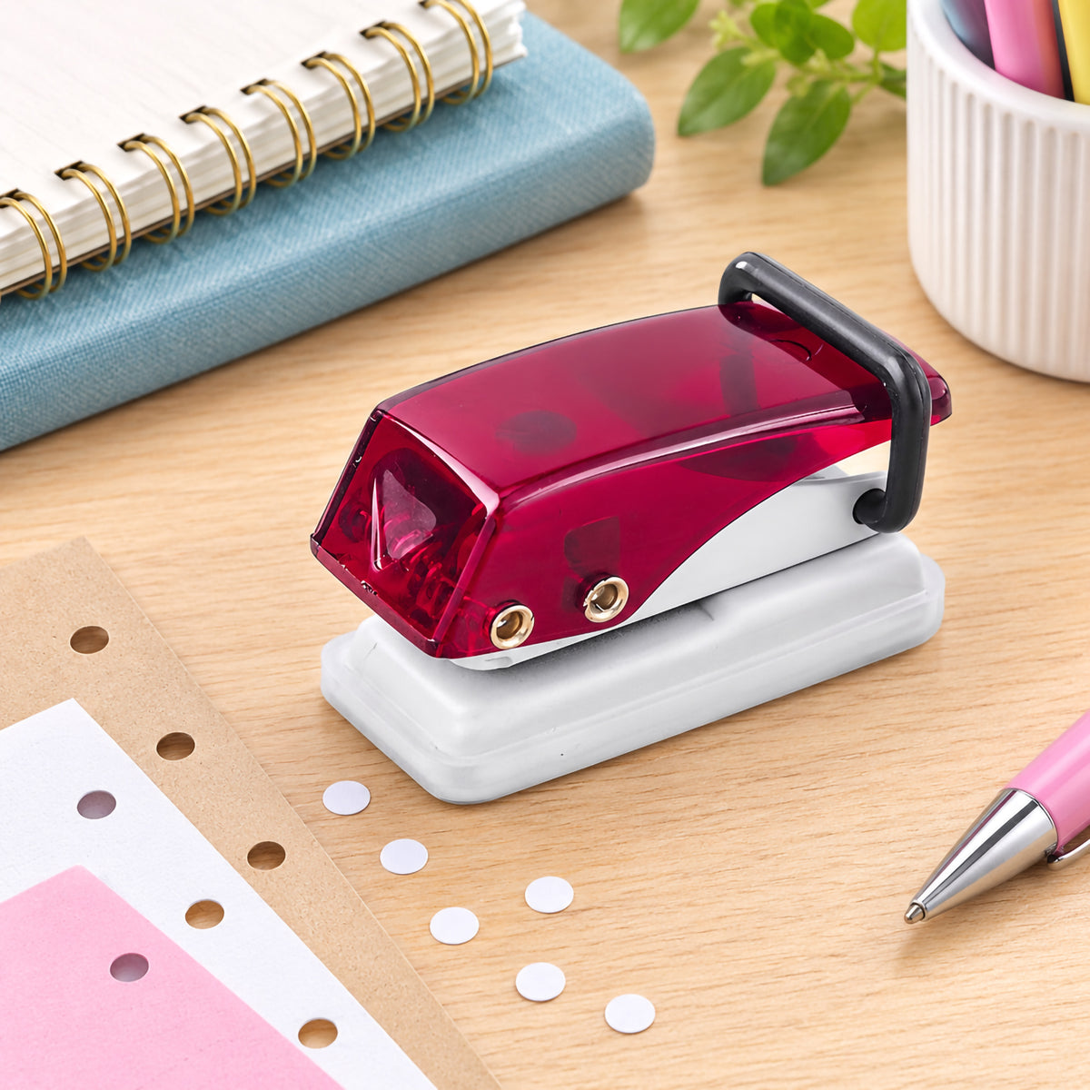 Ruby KW by Colby Little Gem mini paper punch displayed on a timber desk beside punched papers, notebooks and stationery. The compact design features a translucent ruby handle, black rear lever section, metal construction and light coloured protective base, presenting a practical single hole punch for office, school and creative organisation.