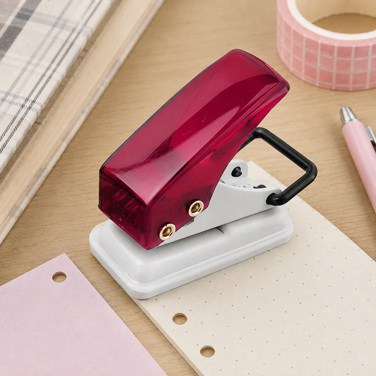 KW by Colby Little Gem ruby one hole punch positioned over dotted paper on a workspace with coloured paper and stationery nearby. The raised handle provides a clear view of the compact hand operated mechanism, sturdy metal body and protective base designed to collect paper confetti after punching.