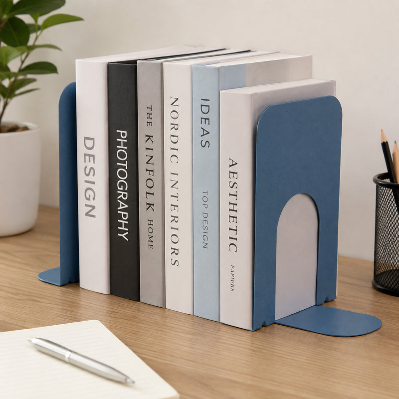 Slate blue KW by Colby student bookends positioned at either end of a row of upright books on a clean office desk. The compact supports hold books of different thicknesses together while their muted blue finish complements a modern workspace with stationery and a notebook nearby.