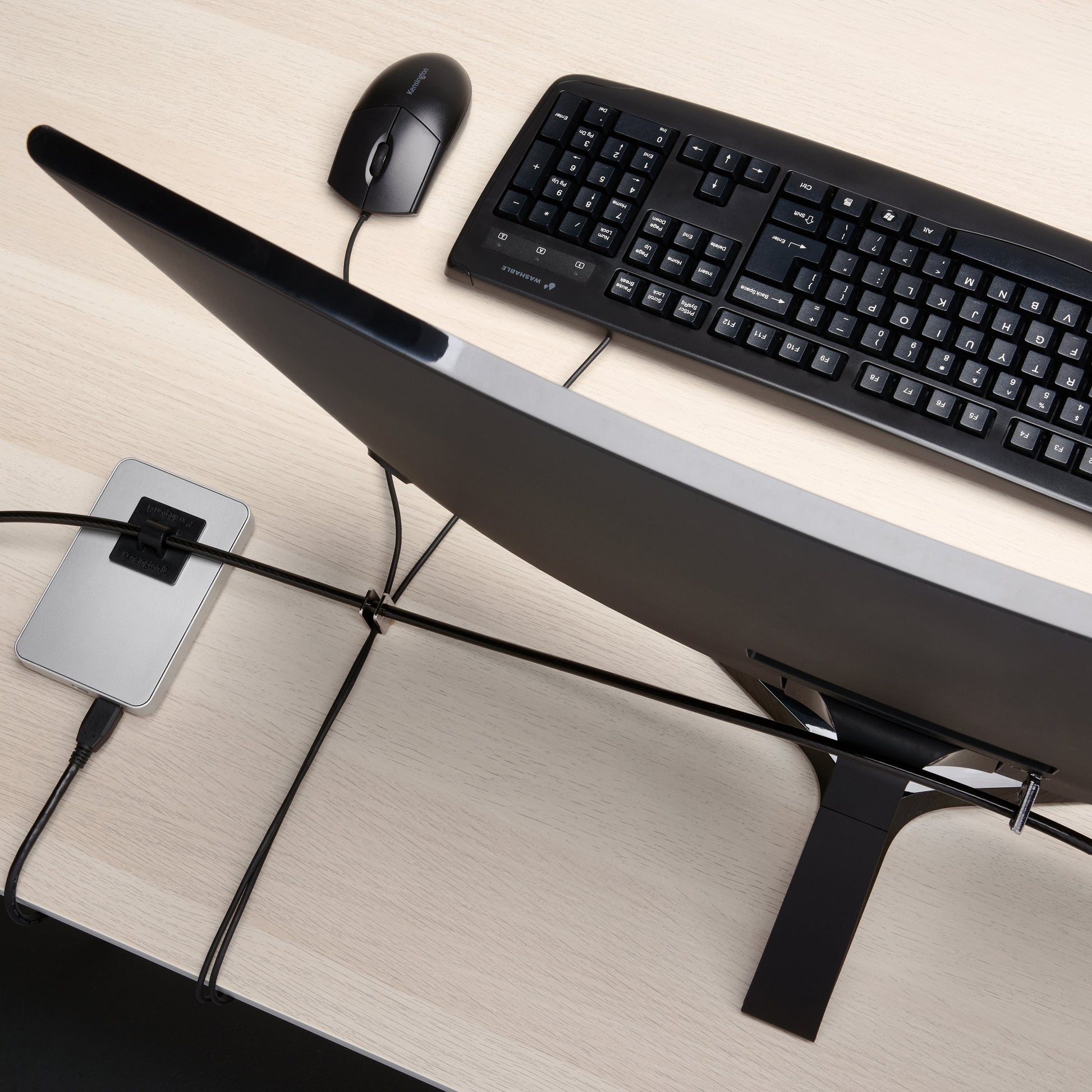 Overhead view of a desktop workstation secured with the Kensington MicroSaver 2.0 T Bar Peripherals Kit. The security cable is routed between a monitor, keyboard, mouse and peripheral device, demonstrating effective protection and cable organisation across multiple desktop accessories.