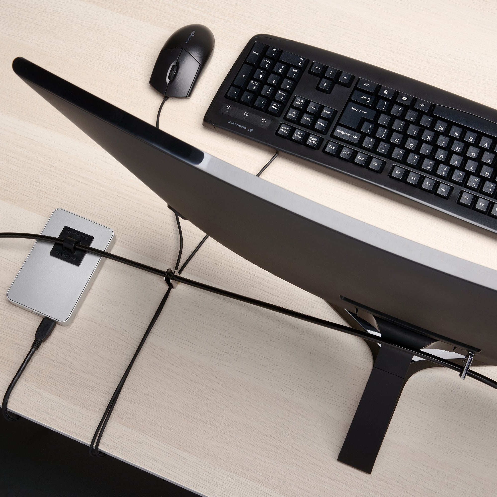 Overhead view of a desktop workstation secured with the Kensington MicroSaver 2.0 T Bar Peripherals Kit. The security cable is routed between a monitor, keyboard, mouse and peripheral device, demonstrating effective protection and cable organisation across multiple desktop accessories.
