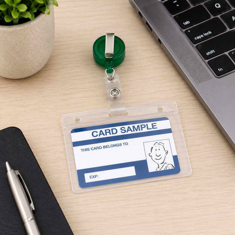 The Kevron ID1013 clear ID card holder with green retractable reel rests on a modern office desk beside a laptop, notebook and pen. A sample identification card is clearly displayed, highlighting the holder's professional appearance and easy card visibility.