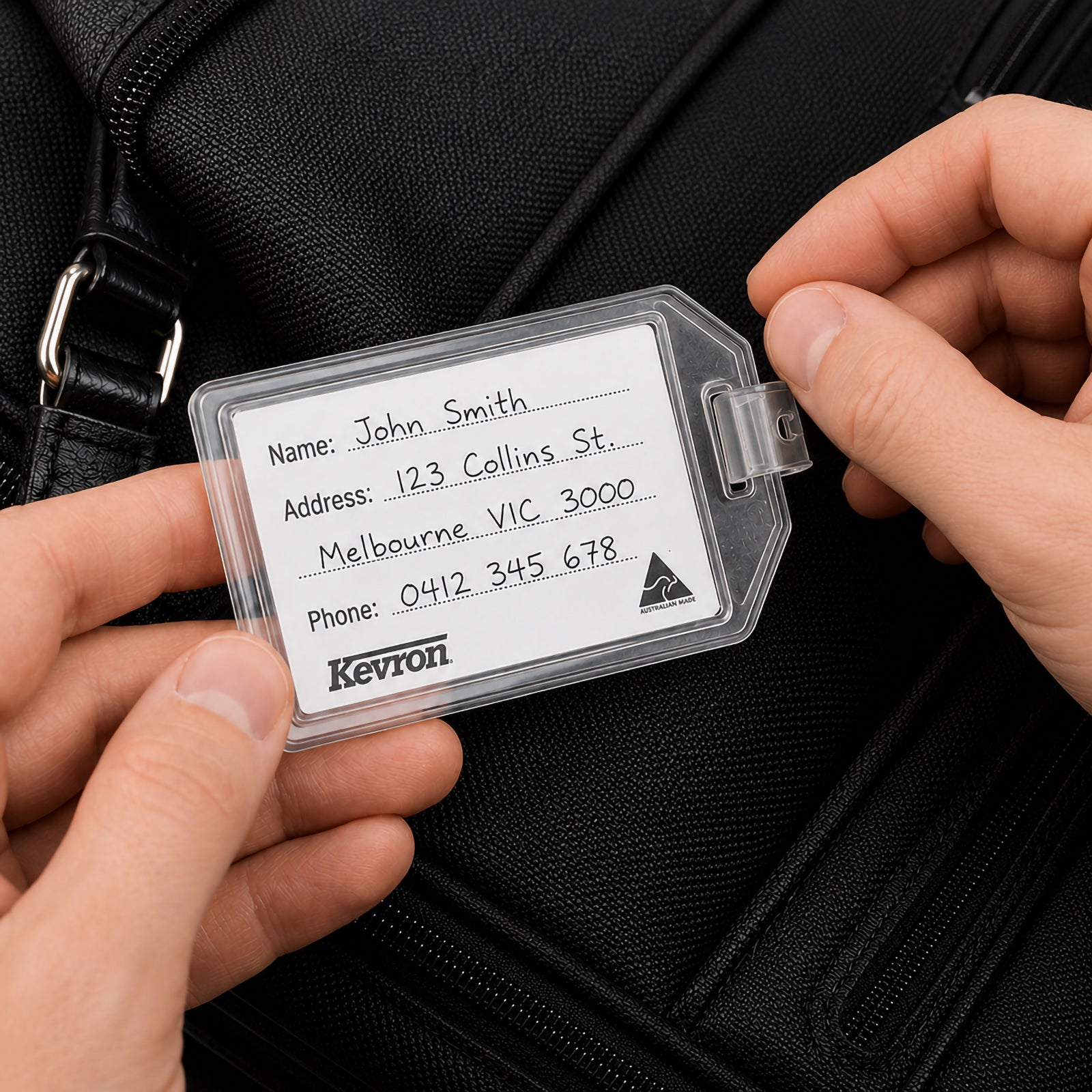 A close up of the Kevron ID34 Executive Hotel Tag being opened while attached to a black travel bag. The user is inserting a personalised identification card, demonstrating the reusable design and secure double lock construction for protecting contact details during travel.
