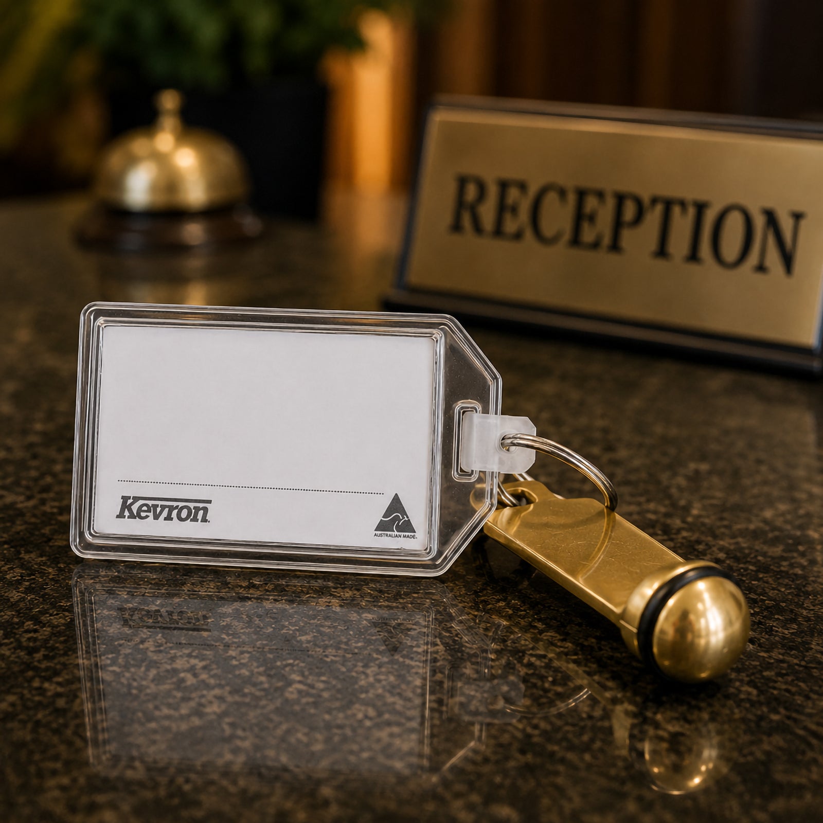 A Kevron ID34 Executive Hotel Tag displayed on a hotel reception counter beside a traditional room key and reception sign. The scene highlights its suitability for hotel guest luggage, accommodation providers and hospitality environments.