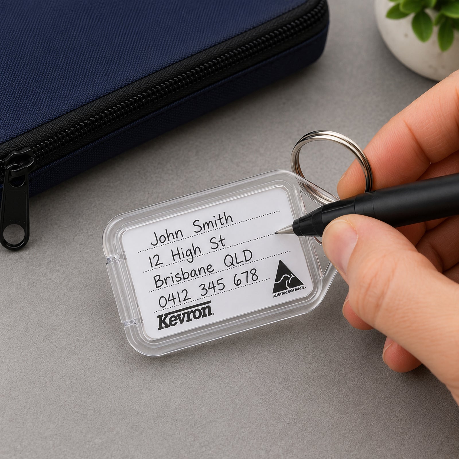 A close up of a Kevron ID5 Clear Key Tag as a user writes identification details onto the removable insert. The transparent Clicktag is displayed beside a zippered case, demonstrating how easily labels can be updated for everyday key organisation.