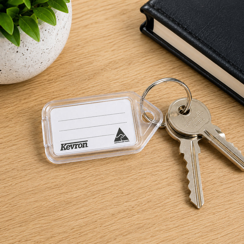 A Kevron ID5 Clear Key Tag attached to a set of household keys on a wooden desk beside a notebook and decorative plant. The scene highlights its suitability for organising home, office and workplace keys.