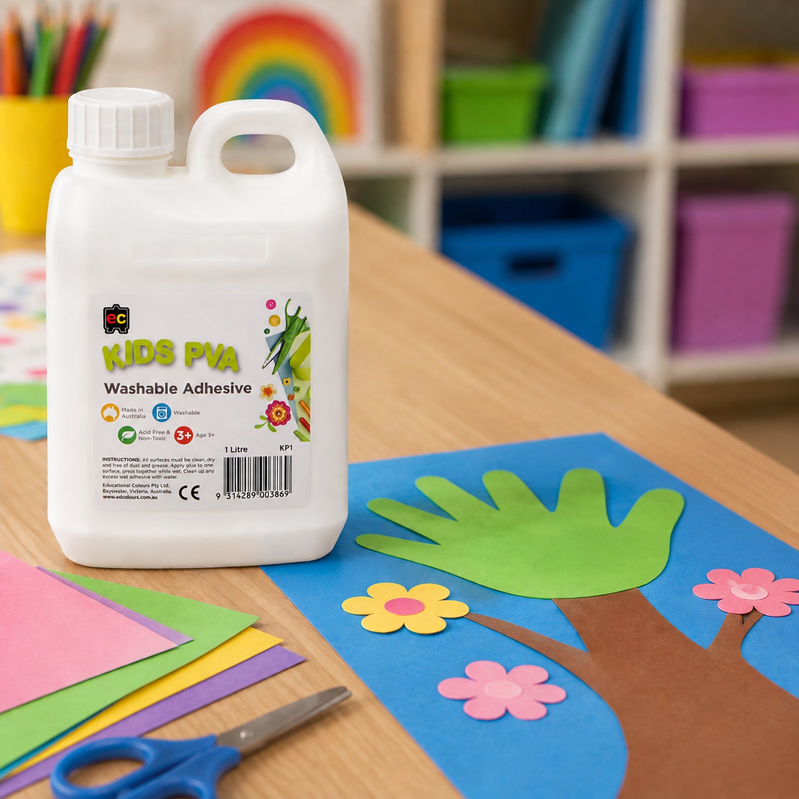 Large 1 litre bottle of Educational Colours Kids Washable PVA Glue positioned next to a handprint tree craft project on a classroom desk. Surrounded by colourful paper sheets and scissors, the image highlights the glue's use for paper crafts, school projects and creative classroom activities.