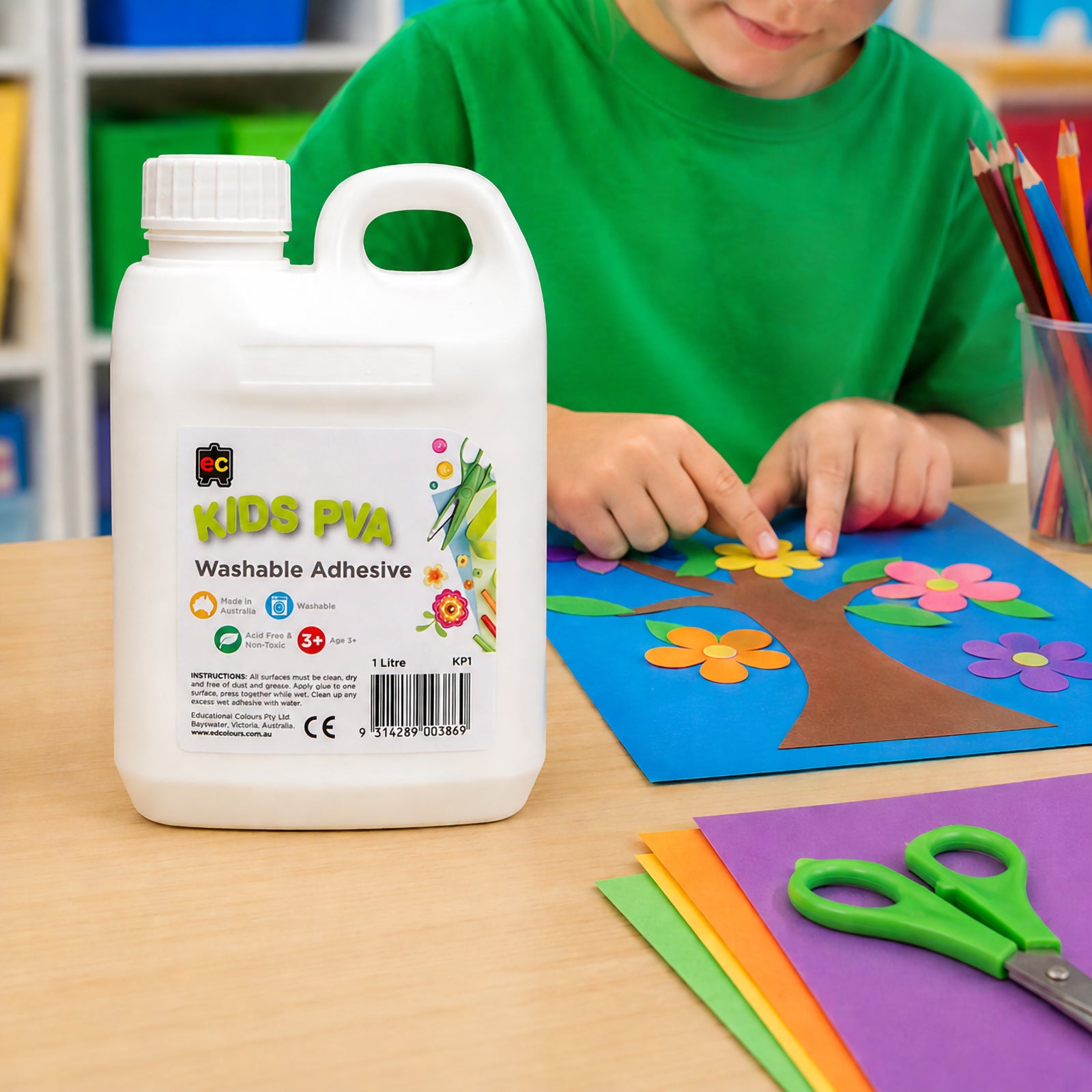 Educational Colours Kids Washable PVA Glue 1 Litre displayed on a classroom table beside a colourful paper flower craft project. A child is actively creating artwork, demonstrating the glue's suitability for school activities, art lessons and hands on learning. Coloured pencils, craft paper and classroom storage units create a bright educational environment.
