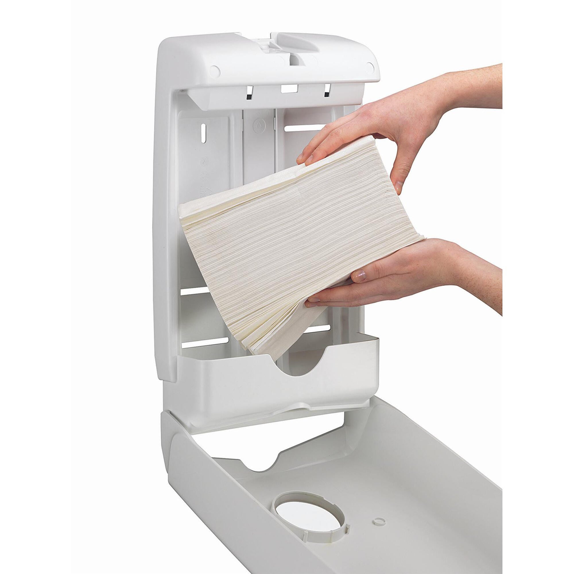 Two hands loading a stack of white Kleenex compact hand towels into an open white Kimberly-Clark dispenser, showing the hinged cover, internal guide plates and the drop down front panel