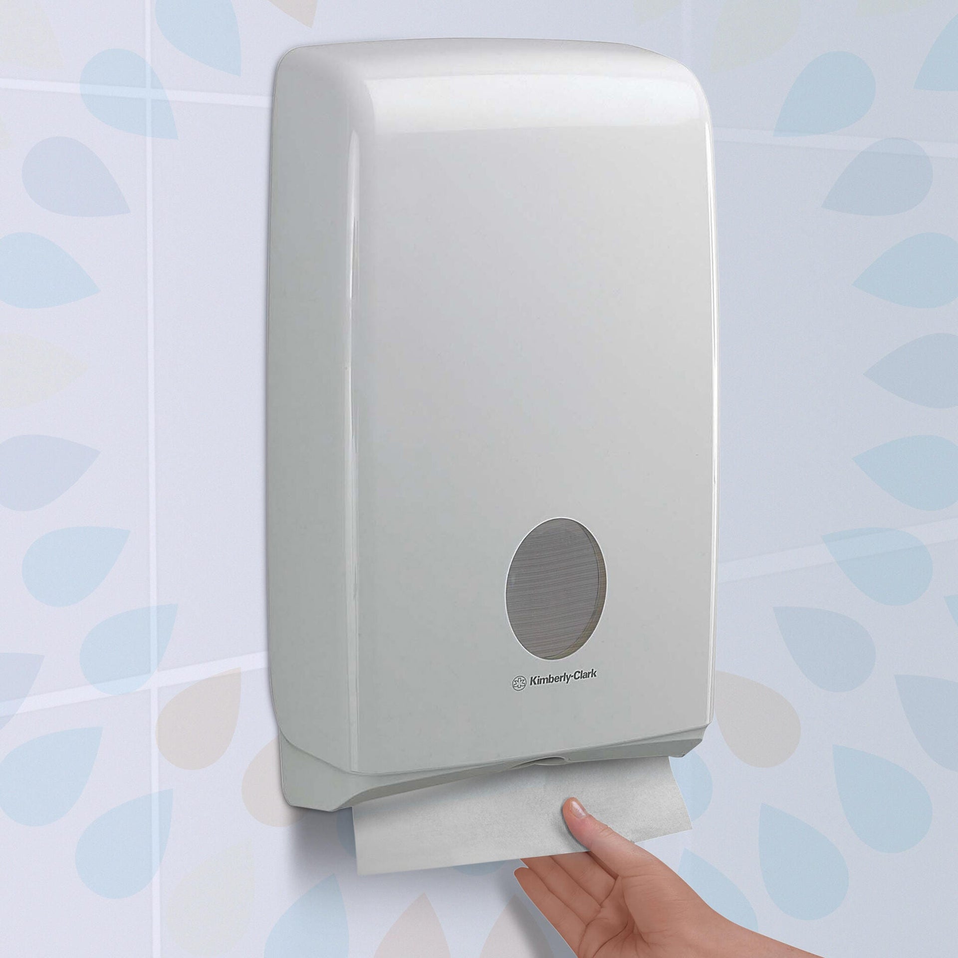 White Kimberly-Clark compact hand towel dispenser mounted on a tiled washroom wall with a hand drawing a single white towel from the slot underneath, showing the oval level window on the cover