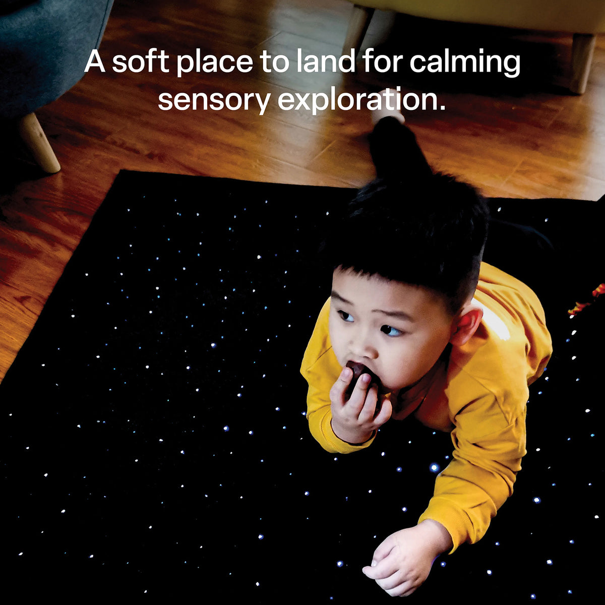 Lifestyle image of a child lying on the illuminated sensory carpet with text describing its use as a soft space for calming sensory exploration. The black carpet displays small blue and white fibre optic lights beneath the child and demonstrates how the product can be incorporated into supervised relaxation and sensory activities.