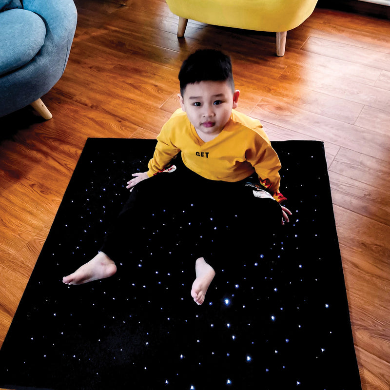 Child seated on the black LED Sensory Carpet during supervised floor based sensory play. Small illuminated fibre optic points create a starry visual effect around the child, demonstrating the generous square surface and how the carpet can provide an engaging area for quiet observation, visual exploration and sensory activities.