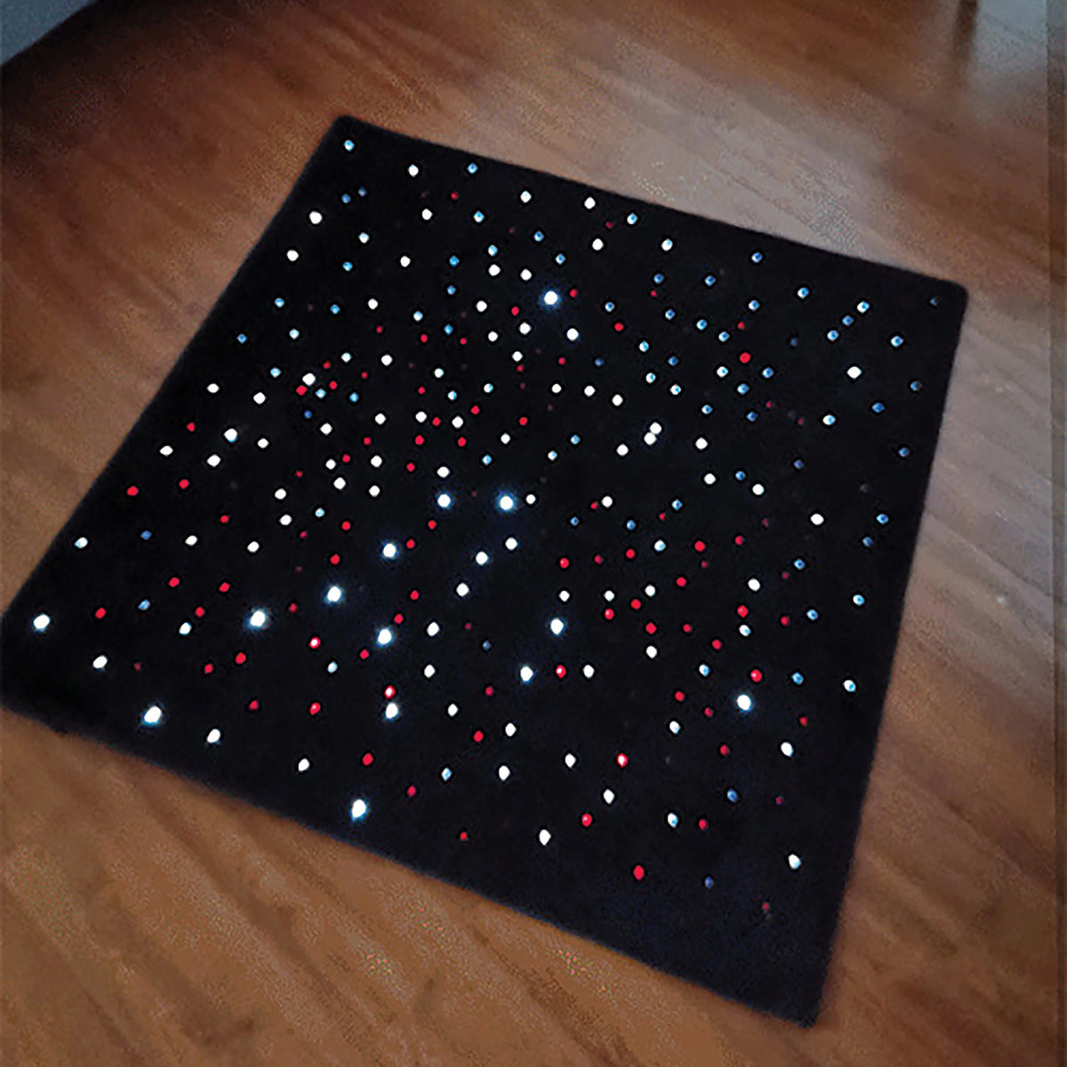 Angled view of the 95 x 95cm LED Sensory Carpet positioned on a timber floor. The black carpet is illuminated with numerous red, blue and white fibre optic points, clearly showing the square shape, soft surface and distributed lighting pattern designed to provide engaging visual sensory stimulation.