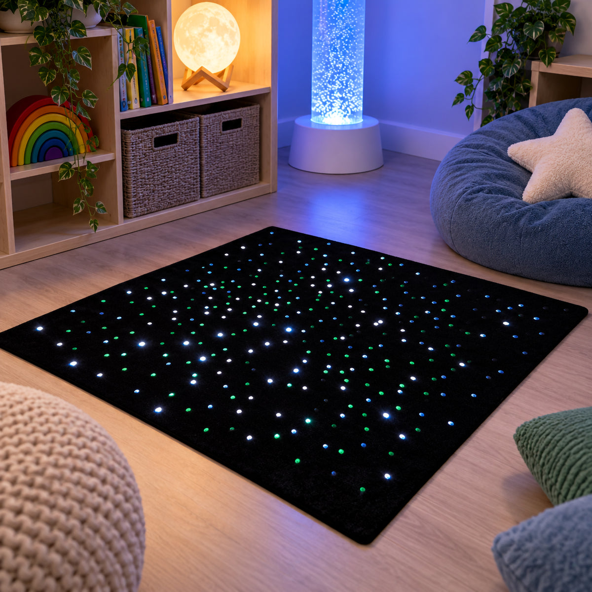 MTA Essentials rechargeable fibre optic sensory carpet displayed in a dedicated sensory room with soft seating, shelving and illuminated sensory equipment. Green, blue and white points of light are distributed across the black carpet surface, showing how the 95 x 95cm carpet can create a defined visual exploration area within a calming environment.