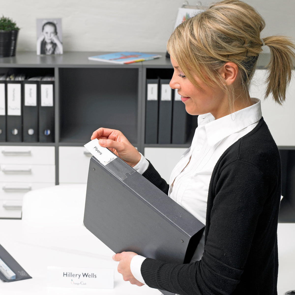 Office worker changing the insert in a 3L label holder on a black ring binder, with labelled binders on shelves behind