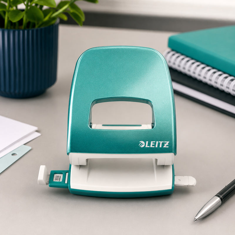 Front view of the Leitz NeXXt WOW 2 Hole Punch in Ice Blue displayed on a contemporary office desk. The punch is shown open with its adjustable paper guide visible, surrounded by a notebook, pen and desktop plant, showcasing its clean design and premium finish.