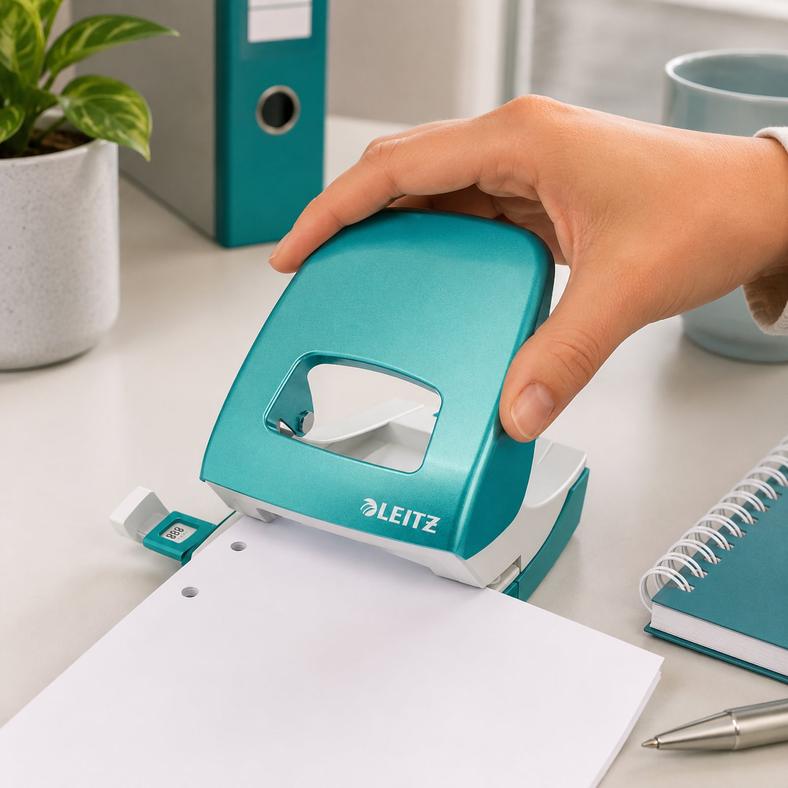 A hand comfortably operates the Leitz NeXXt WOW 2 Hole Punch in Ice Blue while punching a stack of white paper on a modern office desk. The workspace features matching stationery, a teal binder, notebook, pen and indoor plant, highlighting the punch's ergonomic design and effortless everyday performance.