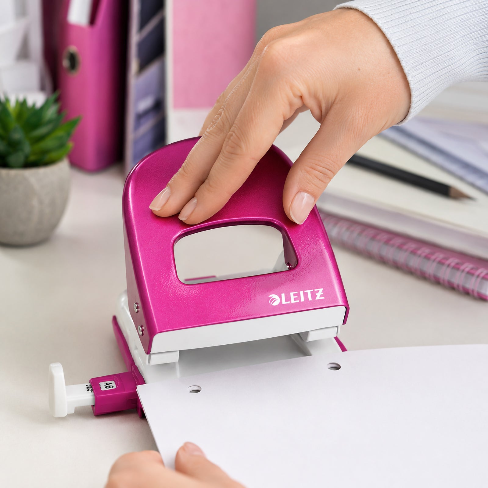 A hand operates the Leitz NeXXt WOW 2 Hole Punch in vibrant Pink while punching white paper on a stylish office desk. Pink folders, notebooks and stationery create a coordinated workspace that highlights the punch's ergonomic grip, smooth operation and colourful professional design.