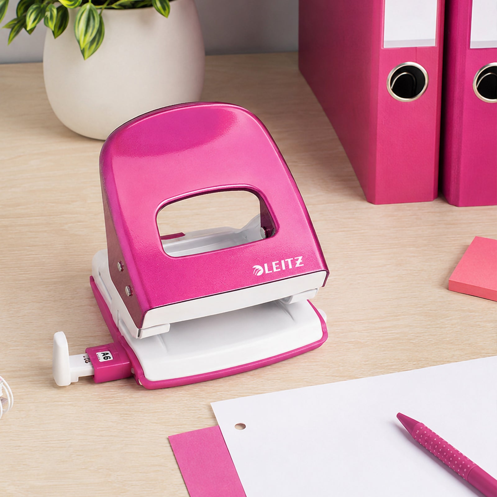 The Leitz NeXXt WOW 2 Hole Punch in Pink sits on a tidy wooden office desk surrounded by matching binders, writing accessories and paperwork. The lifestyle setting emphasises its durable construction, vibrant appearance and suitability for everyday office organisation.