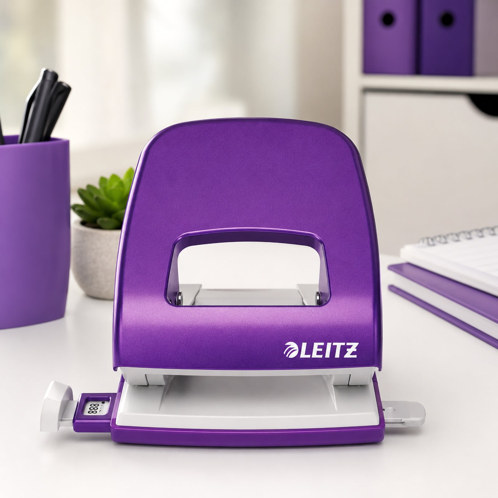 Front facing view of the Leitz NeXXt WOW 2 Hole Punch in Purple displayed on a clean office desk. The punch is shown open with its adjustable paper guide visible, surrounded by matching purple stationery, a pen holder and notebooks that emphasise its premium finish and professional appearance.