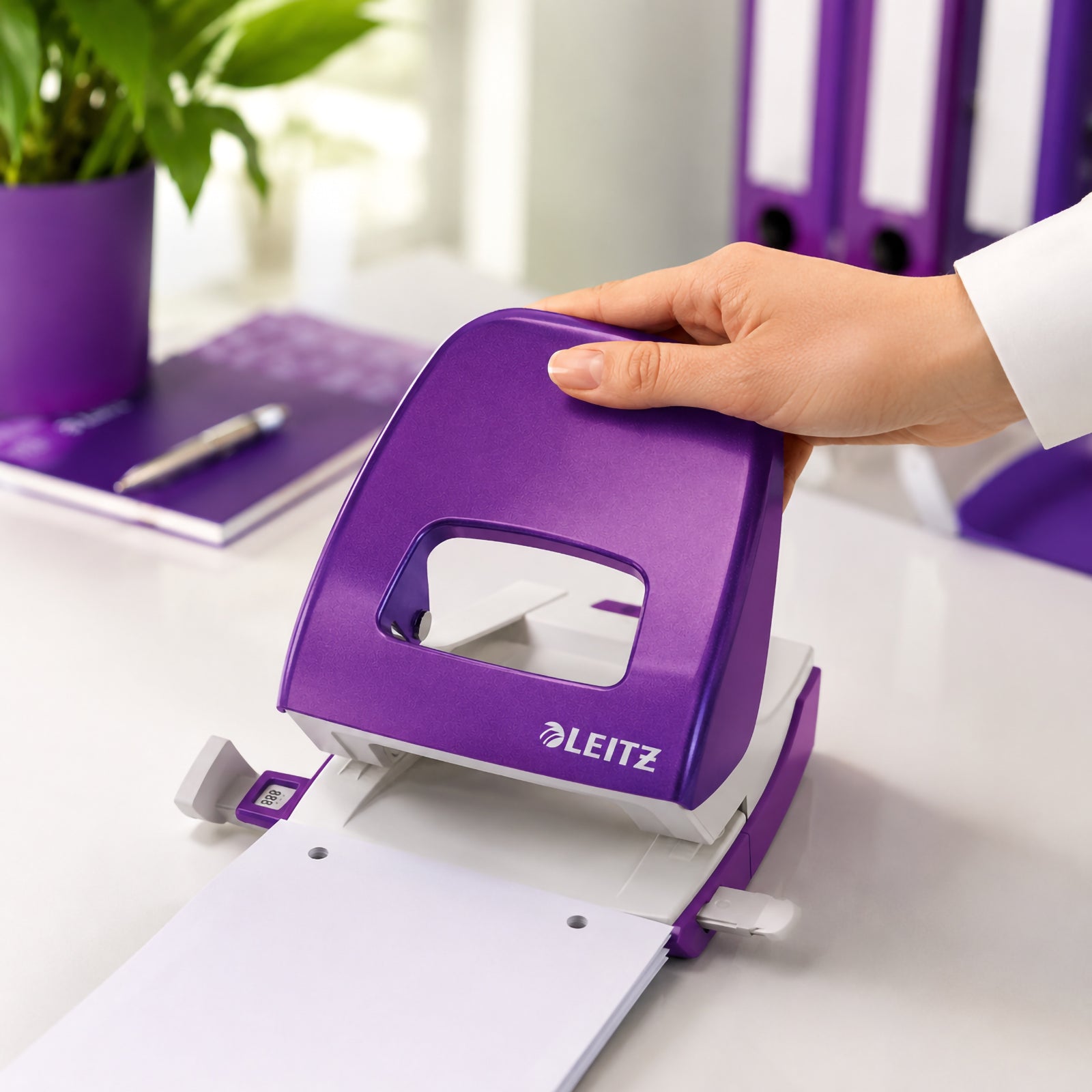 A hand presses the Leitz NeXXt WOW 2 Hole Punch in vibrant Purple while punching white paper on a modern office desk. Purple binders, a matching notebook and a desktop plant create a coordinated workspace that highlights the punch's ergonomic design, smooth operation and contemporary office style.