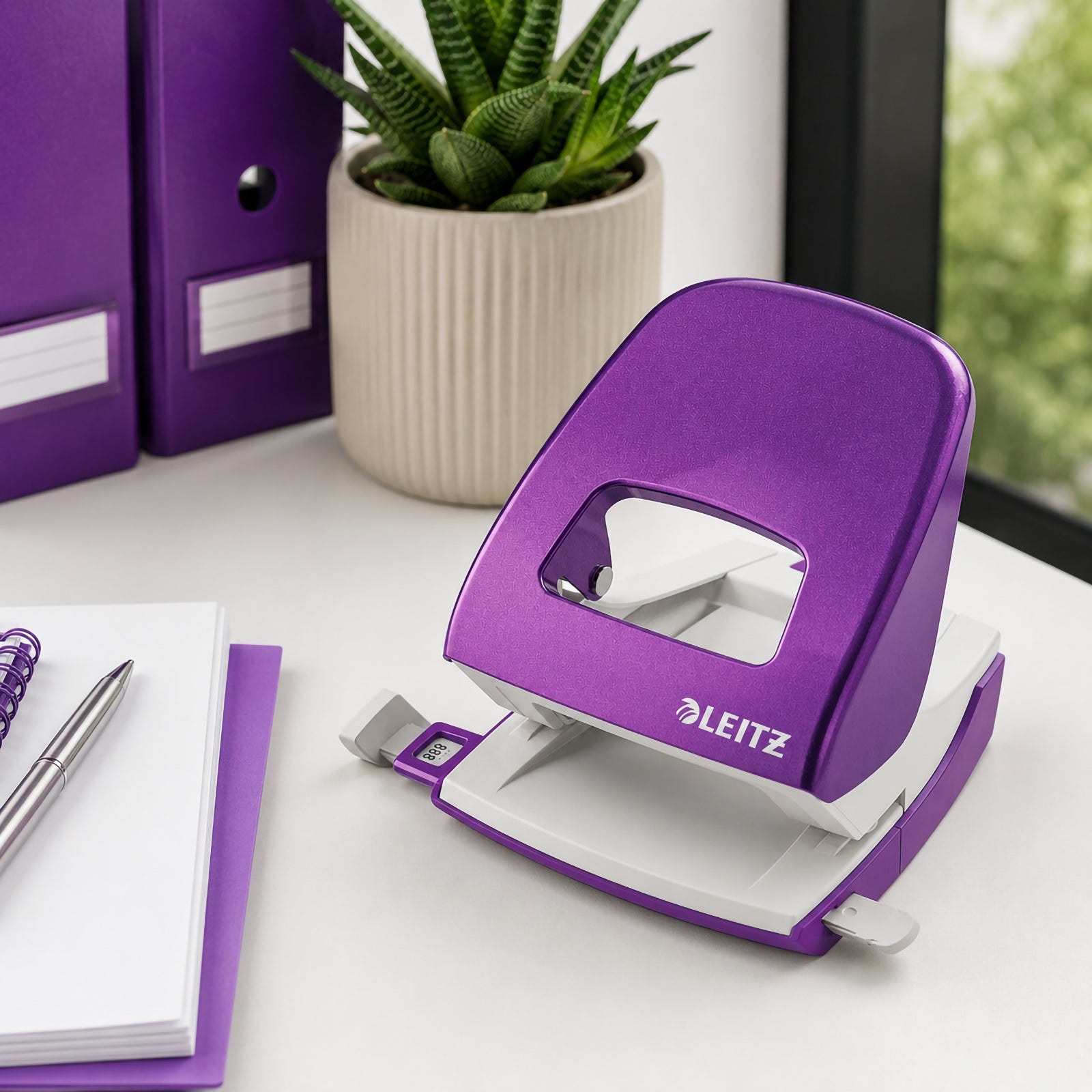 The Leitz NeXXt WOW 2 Hole Punch in Purple sits on a bright office desk beside organised binders, a notebook, pen and decorative succulent. The angled lifestyle scene showcases the punch's modern design, vibrant colour and practical functionality within a productive workspace.
