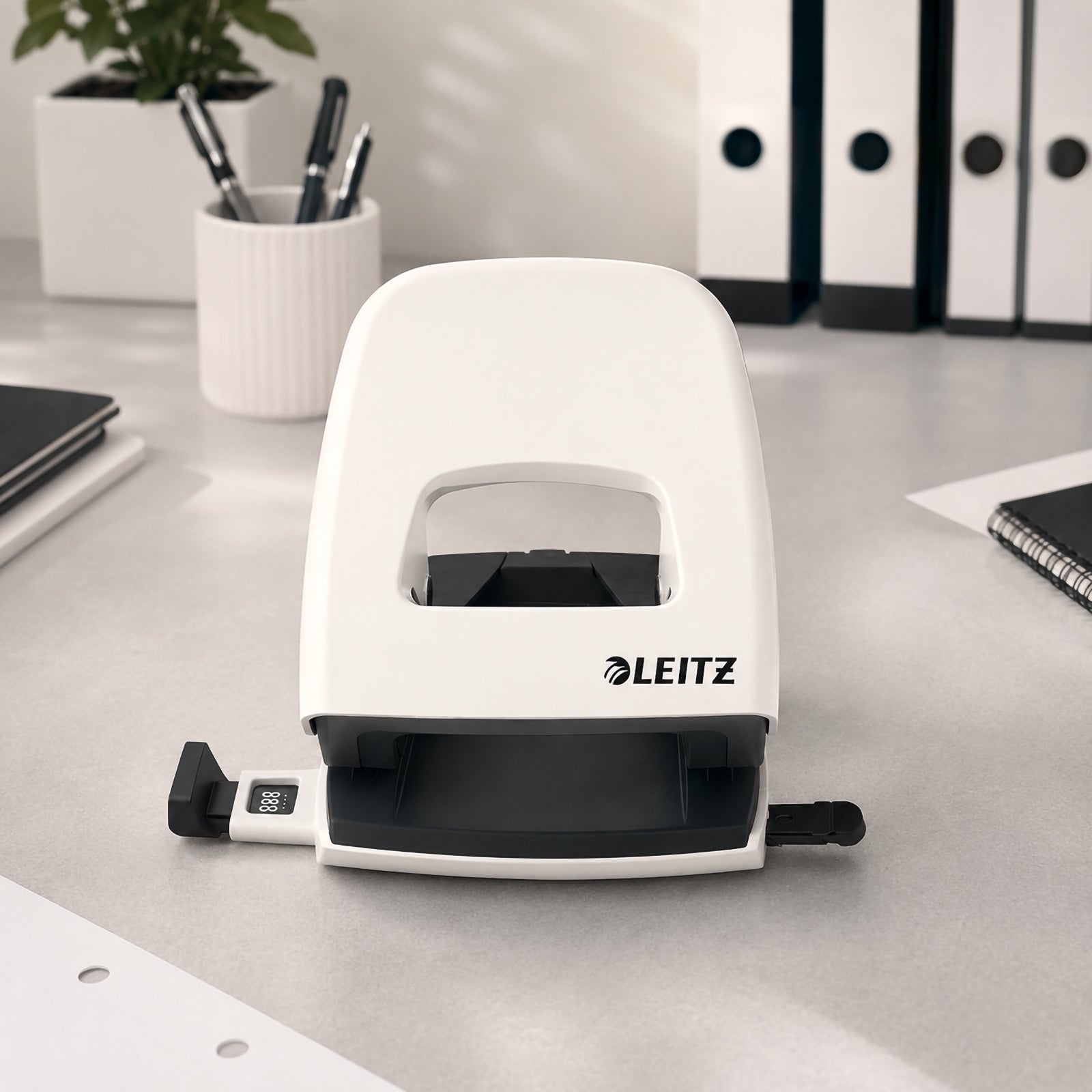 Front view of the Leitz NeXXt WOW 2 Hole Punch in White displayed on a tidy office desk. The punch is shown open with its adjustable paper guide visible, surrounded by office folders, pens and a desktop plant, emphasising its minimalist design and premium finish.