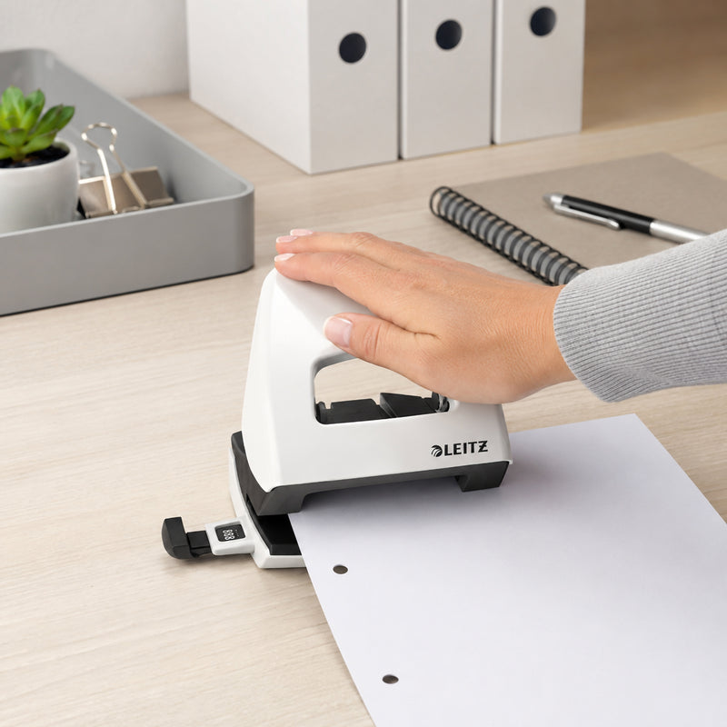 A hand operates the Leitz NeXXt WOW 2 Hole Punch in White while punching sheets of paper on a modern office desk. White storage boxes, a notebook and desktop organiser create a clean professional workspace that highlights the punch's ergonomic operation, comfortable grip and reliable everyday performance.