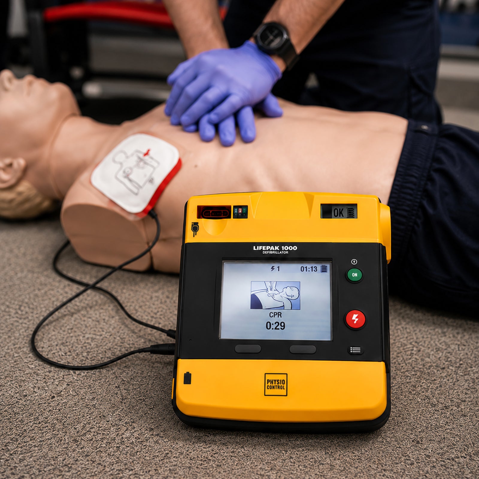 LifePak 1000 Semi Automatic Defibrillator positioned beside a CPR training manikin while a responder performs chest compressions. The connected electrode pad and illuminated display demonstrate realistic emergency response training using advanced CPR guidance.