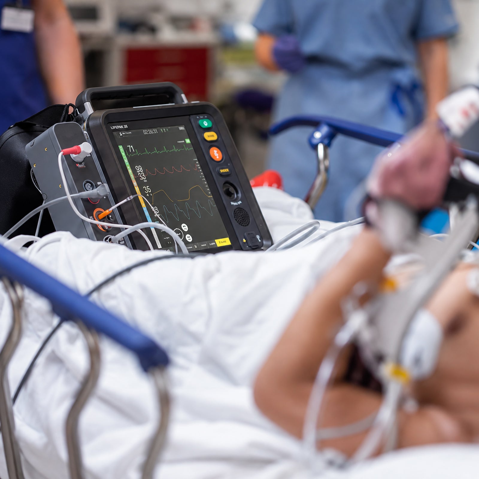 LifePak 35 Defibrillator Monitor positioned beside a hospital patient during acute clinical care. Multiple monitoring waveforms are visible on the colour display while medical staff work around the patient, illustrating continuous cardiac and vital signs assessment during a busy emergency treatment workflow.