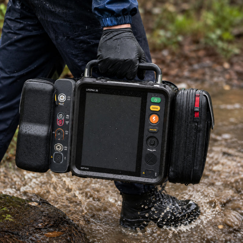 LifePak 35 Defibrillator Monitor being carried by its integrated handle through a wet outdoor environment. Water droplets cover the rugged housing while the large front display, defibrillation controls, monitoring connections and protective accessory compartments remain clearly visible, demonstrating the portable design for demanding emergency response conditions.