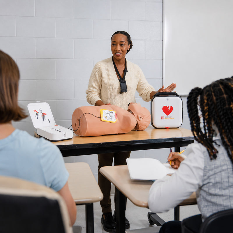 An instructor demonstrates the LifePak CR2 Fully Automatic Defibrillator during a CPR and AED training session using a training manikin. Students observe the lesson as the device and its carry case are displayed on the classroom table, highlighting workplace emergency preparedness.
