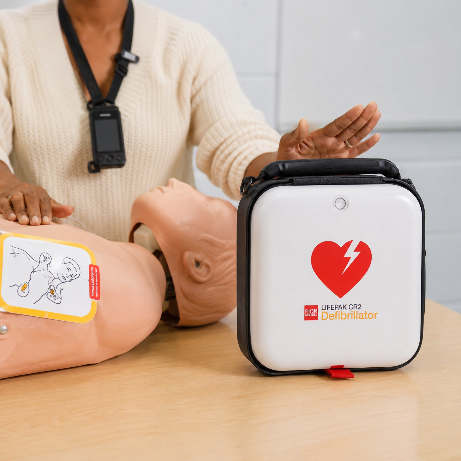 A CPR instructor demonstrates the LifePak CR2 Semi Automatic Defibrillator beside a training manikin during a classroom first aid session. The protective carry case is displayed prominently while students learn correct AED operation and emergency response techniques.
