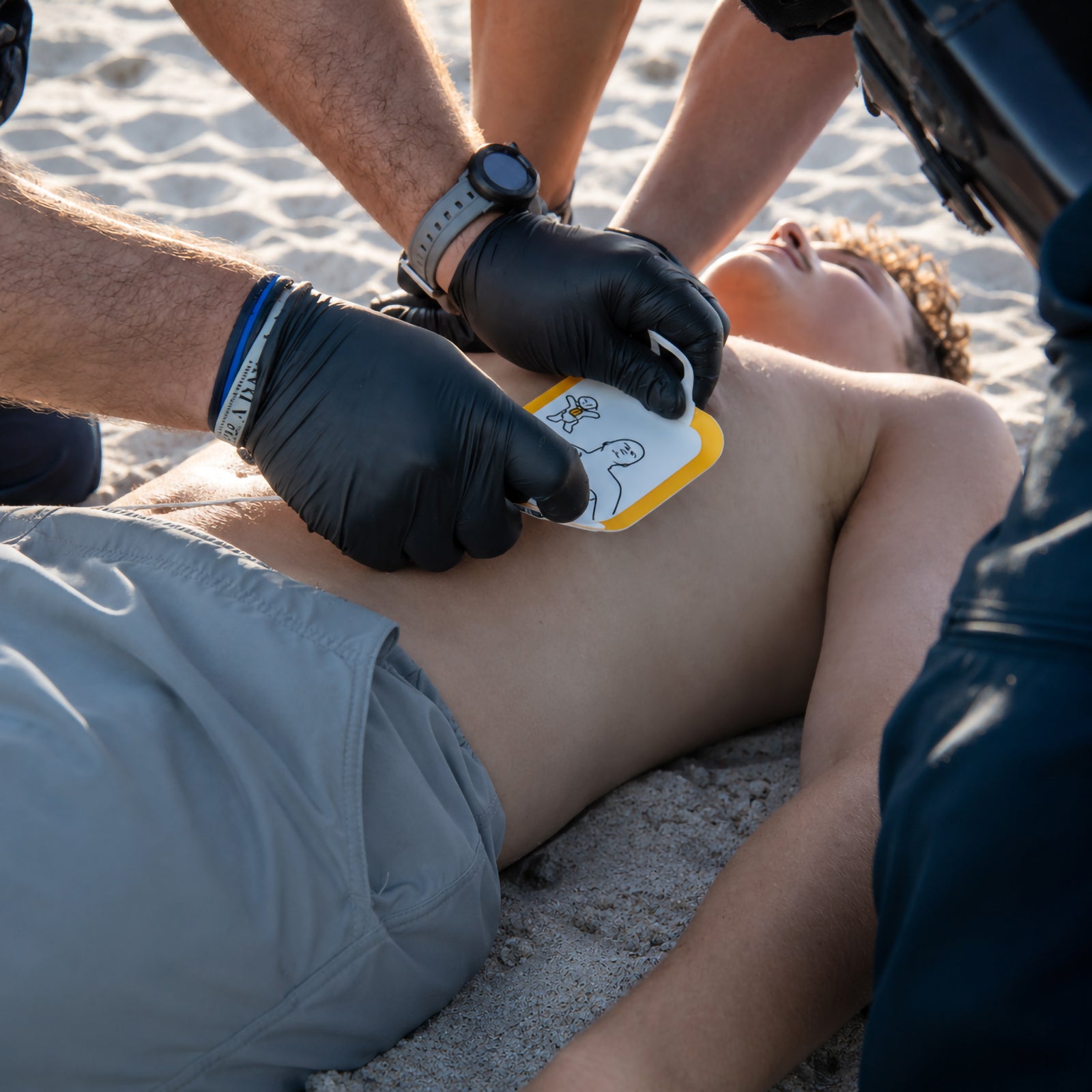 Emergency responders apply a LifePak CR2 electrode pad to a casualty on a sandy beach during a simulated rescue. The close up image demonstrates rapid pad placement in an outdoor emergency, highlighting the device's suitability for public access and first responder situations.