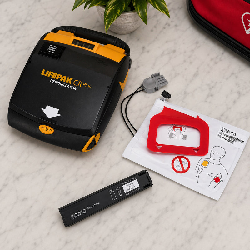 LifePak CR Plus Charge Pak arranged beside a LifePak CR Plus defibrillator in a clean medical lifestyle setting. The adult electrode pad package, connector and battery pack are clearly visible alongside the compatible AED, demonstrating the replacement components used to help maintain the device for emergency response readiness.